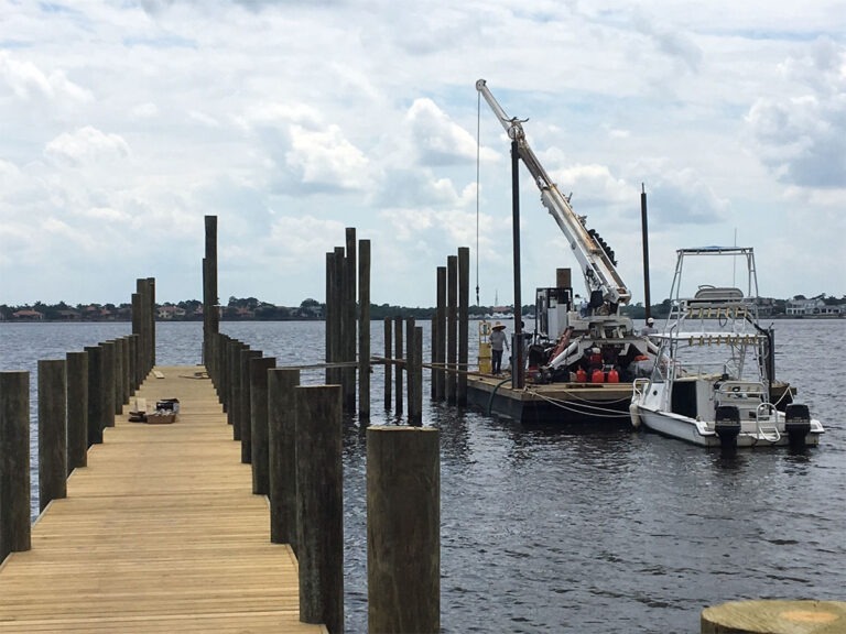 Marine construction crew working from a barge with a crane, installing or repairing wooden pilings on a long dock extending into the water